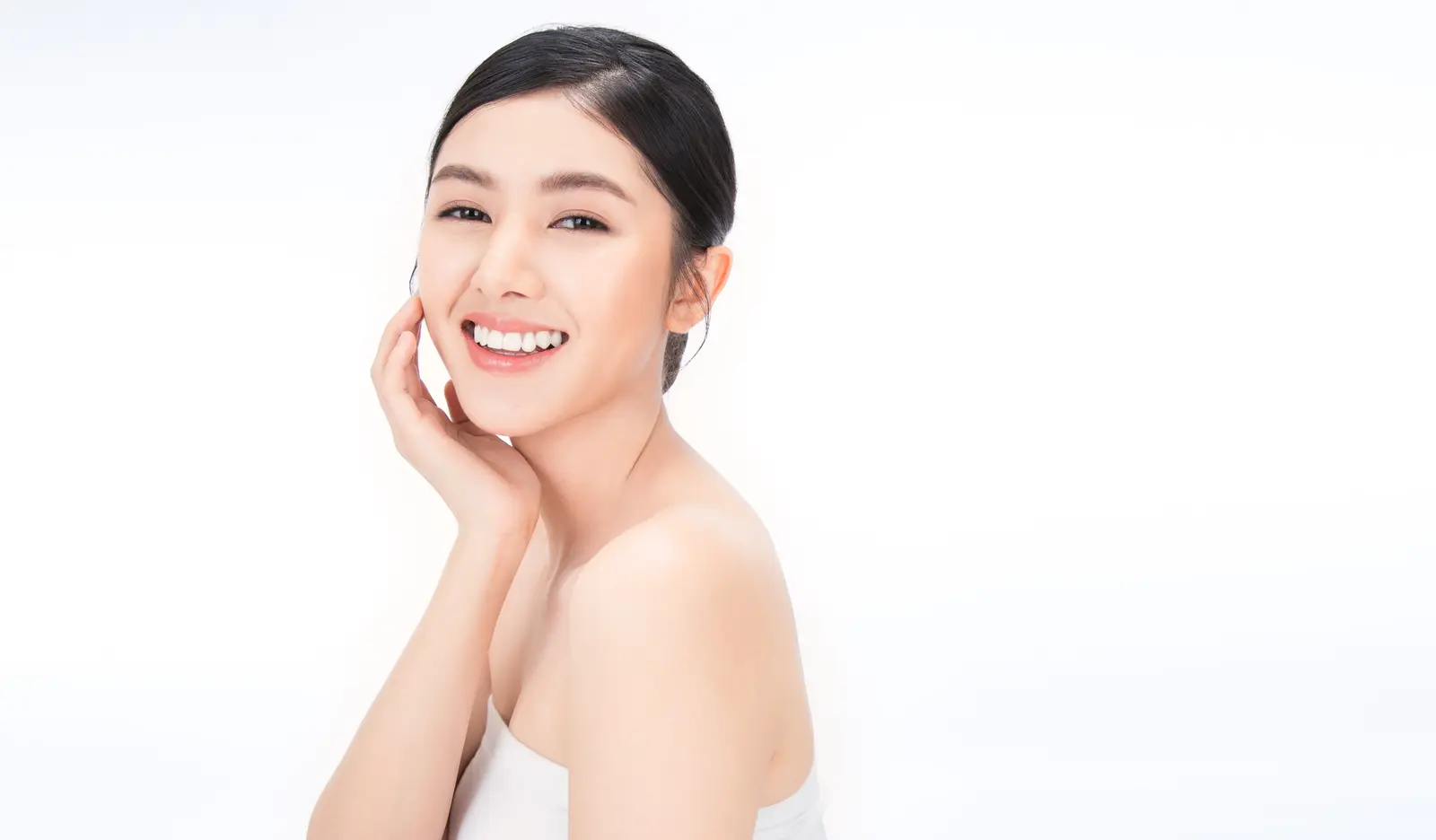 Smiling woman with glowing skin, touching her face against a white background.